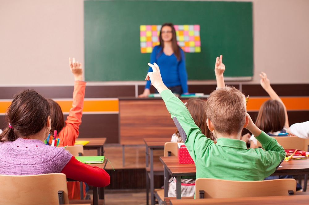 Children In Elementary School Are Raised Hand In Clasroom. Teacher Is In The Background In Front Of Chalkboard.